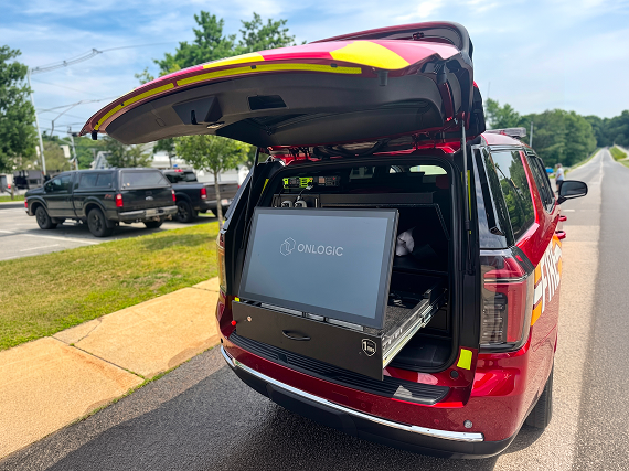 Rear cargo area of a red East Providence Fire SUV, hatch open, showing an OnLogic rack-mounted touchscreen display and equipment drawer installed inside; parked SUVs and trees visible in the background.